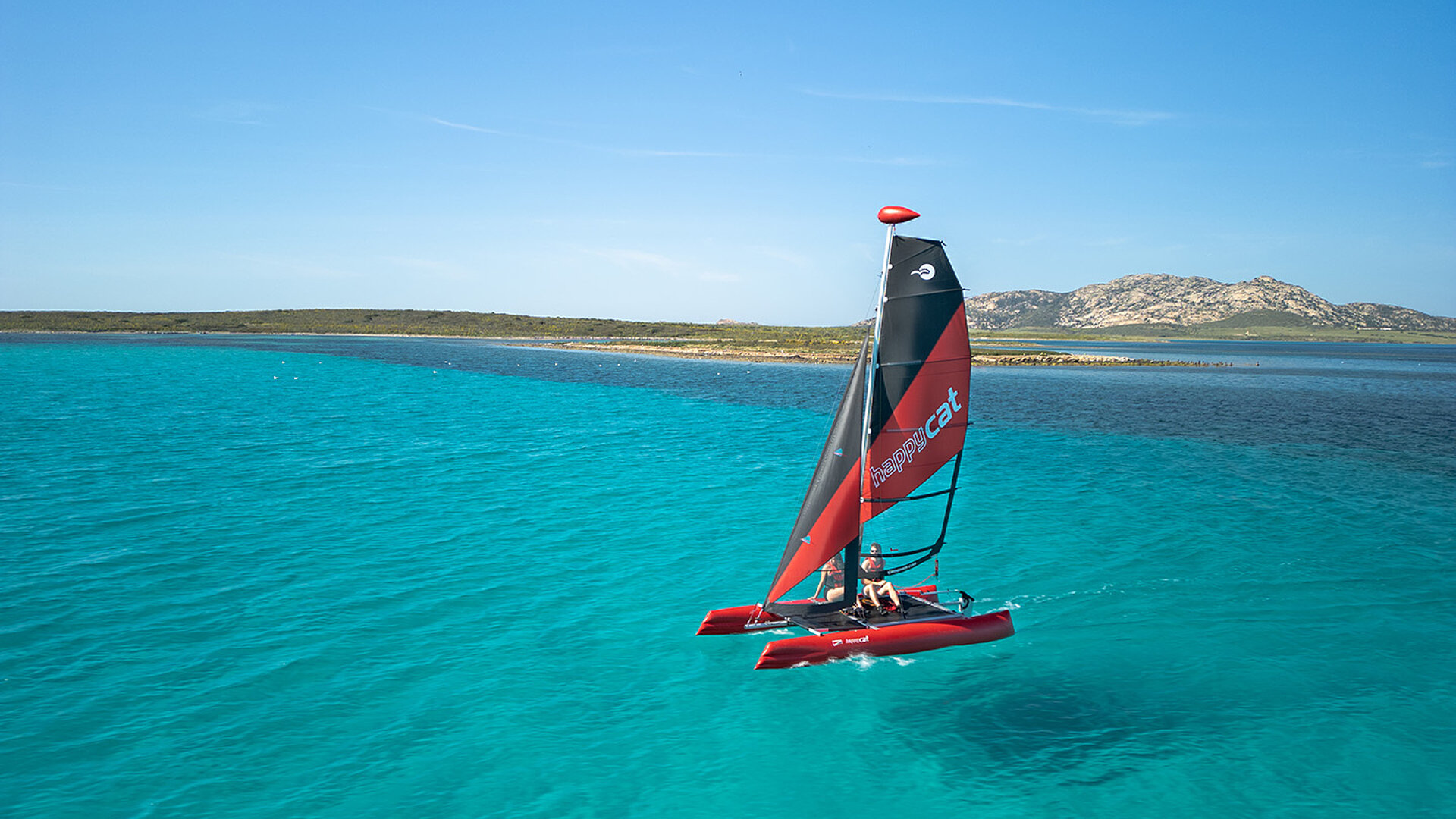 Katamaran Happy Cat Star mit rot-schwarzem Segel segelt auf klarem blauem Wasser vor Küste mit Hügeln