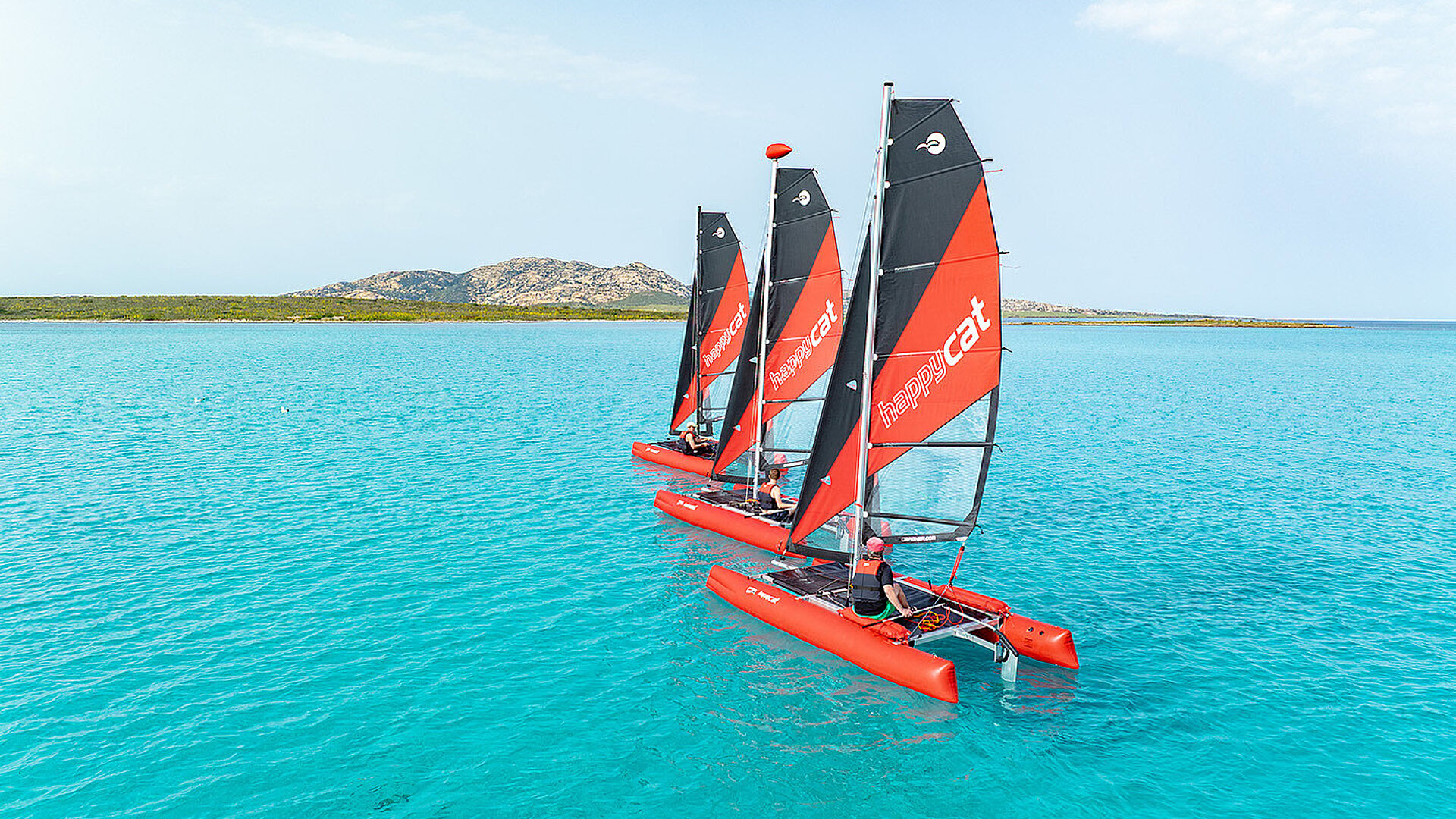 Drei rote und schwarze Happy Cat Star Katamarane segeln auf klarem blauem Wasser mit einer felsigen Küste im Hintergrund